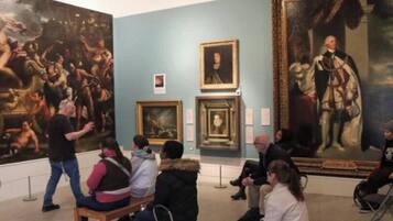 A museum interior with large paintings, a seated audience, and a guide.