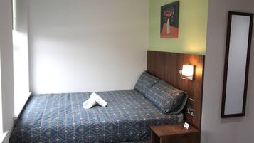Una cama individual con colcha azul y dorada estampada, un cabecero de madera, una mesita de noche con lámpara y un espejo instalado en la pared.