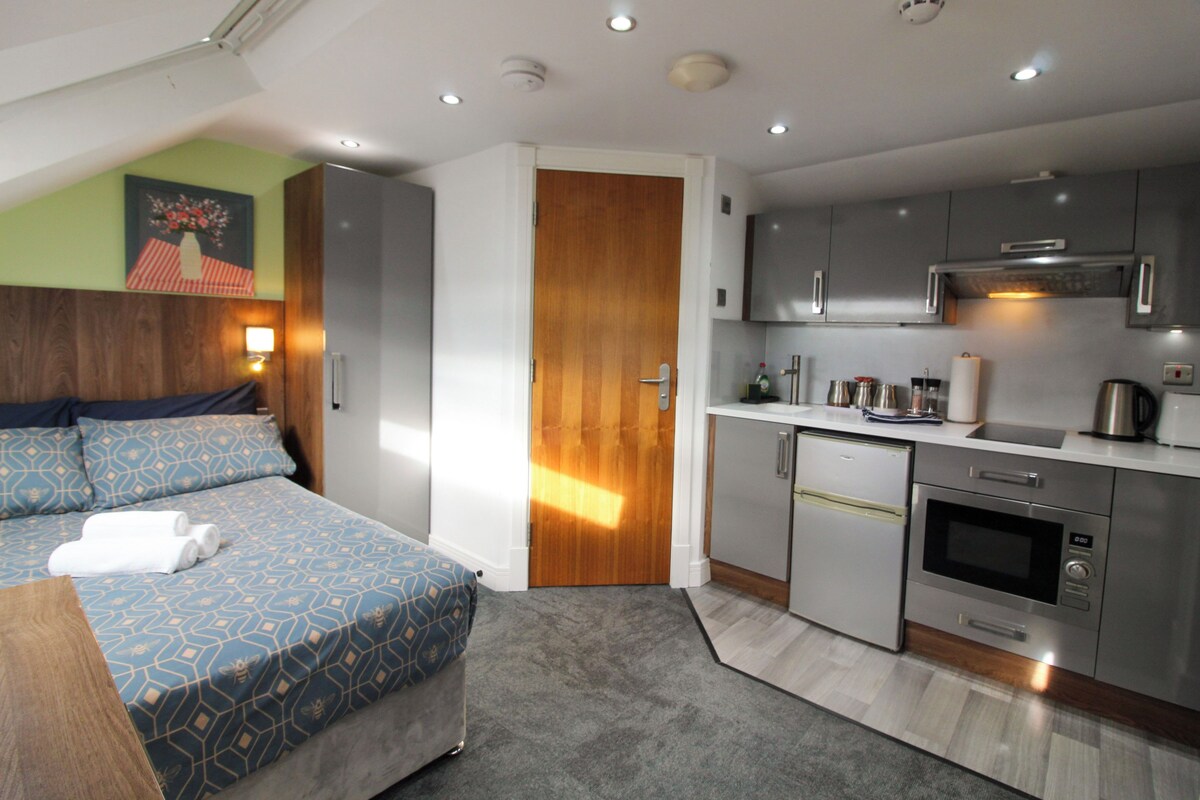 A modern bedroom with a bed, bedside table, and a small kitchenette with a microwave, oven, and sink.
