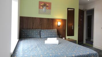 Una cama bien hecha con una colcha de diseño geométrico, una mesita de noche con una lámpara y una puerta a otra habitación.