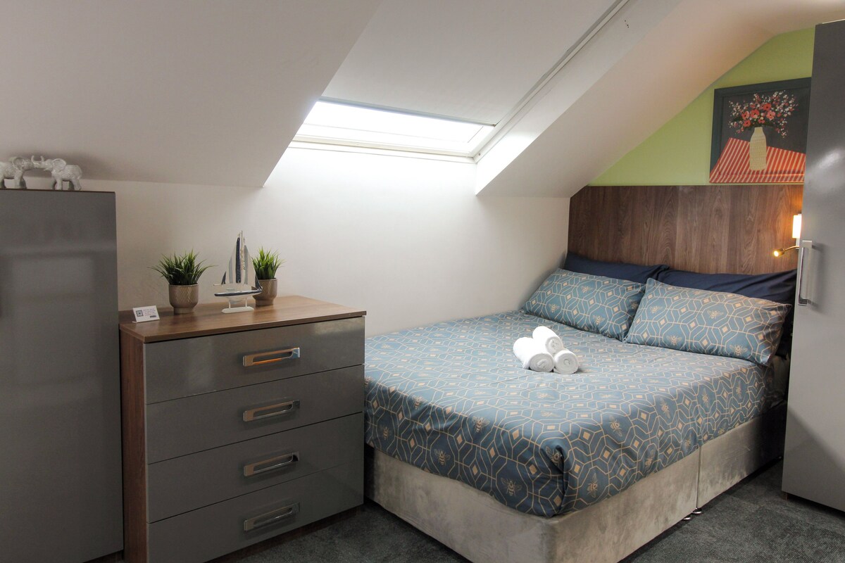 A bedroom with a sloped ceiling, a bed with patterned bedding, a dresser with drawers, and a small potted plant.