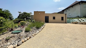 A modern building with a gravel driveway leading to a door, surrounded by a garden with various plants and a small pond.