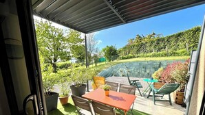 An outdoor patio with a table and chairs, surrounded by potted plants and a view of a garden and a swimming pool.