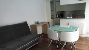 A modern living room with a black sofa, a glass-top dining table with two chairs, a wooden side table, and a kitchen area with a stove, microwave, and cabinets. - Rented T2 independent shared pool at home Near beaches. (Perpignan)