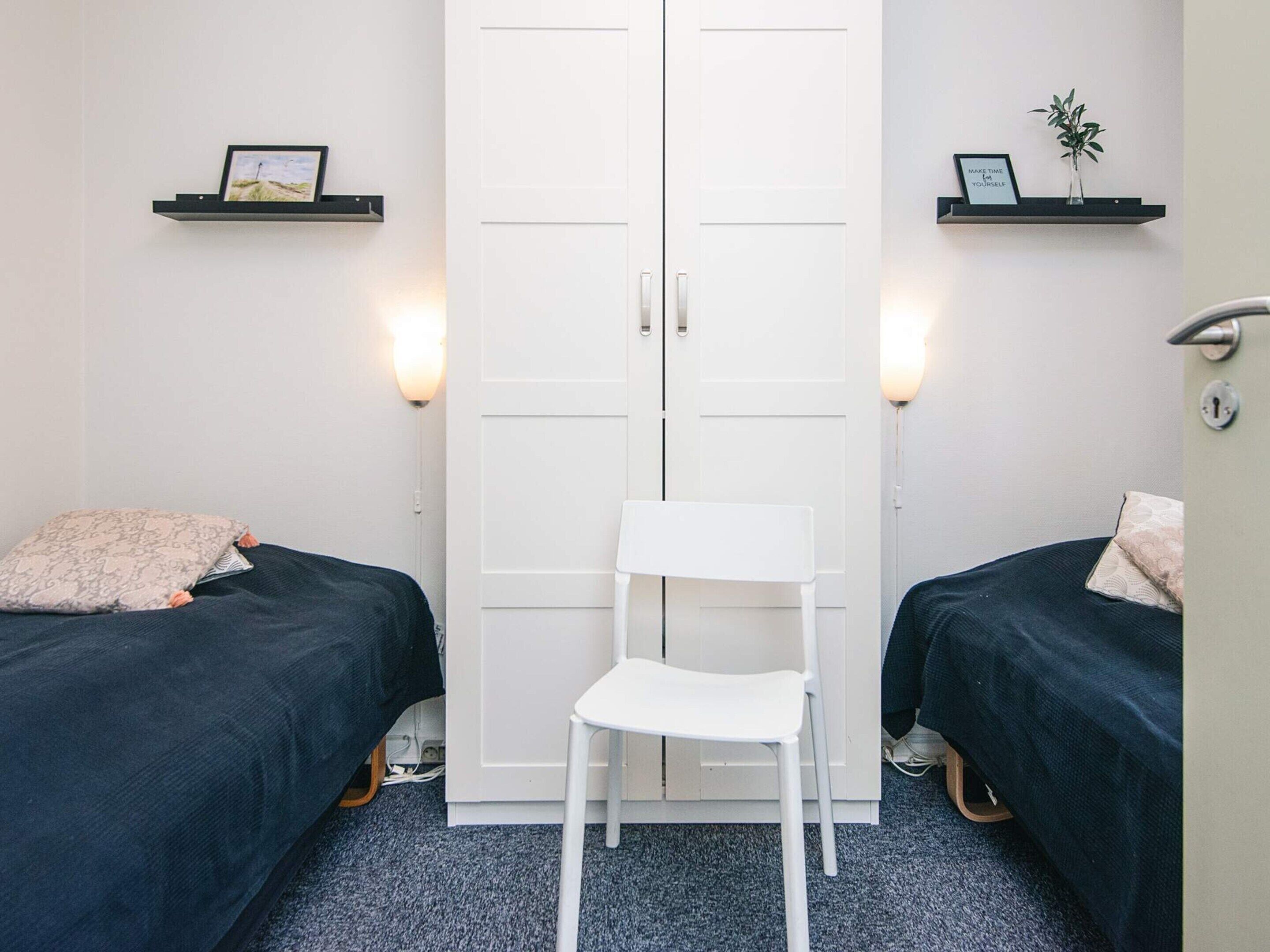 Ein Zimmer mit zwei Betten, einem weißen Stuhl und einem Kleiderschrank. An der Wand sind Regale mit Bildern und einer kleinen Pflanze angebracht.