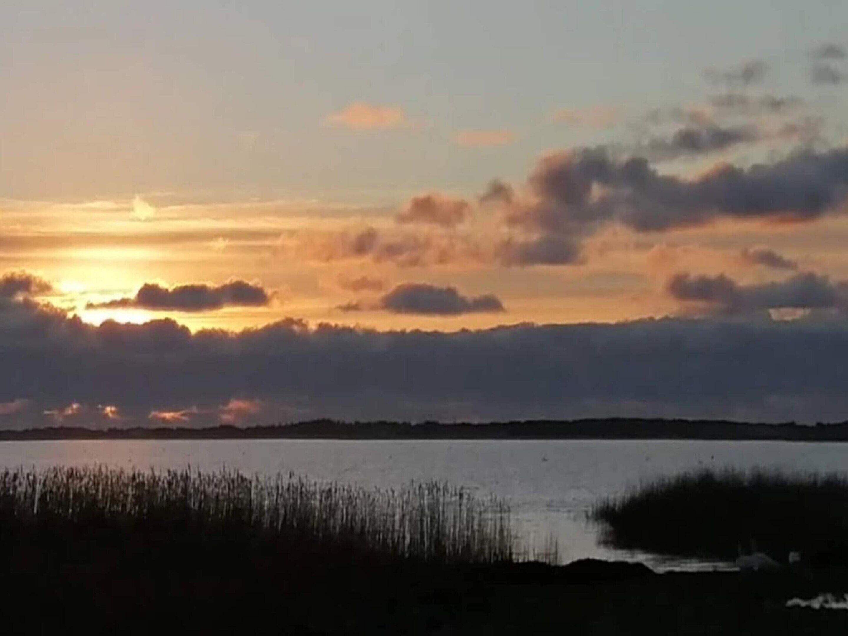 A sunset over a calm body of water with a distant shoreline and overcast sky.