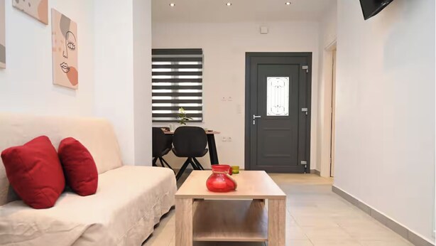 A modern living room with a beige sofa, red cushions, a wooden coffee table with a red vase, and a dark door in the background.