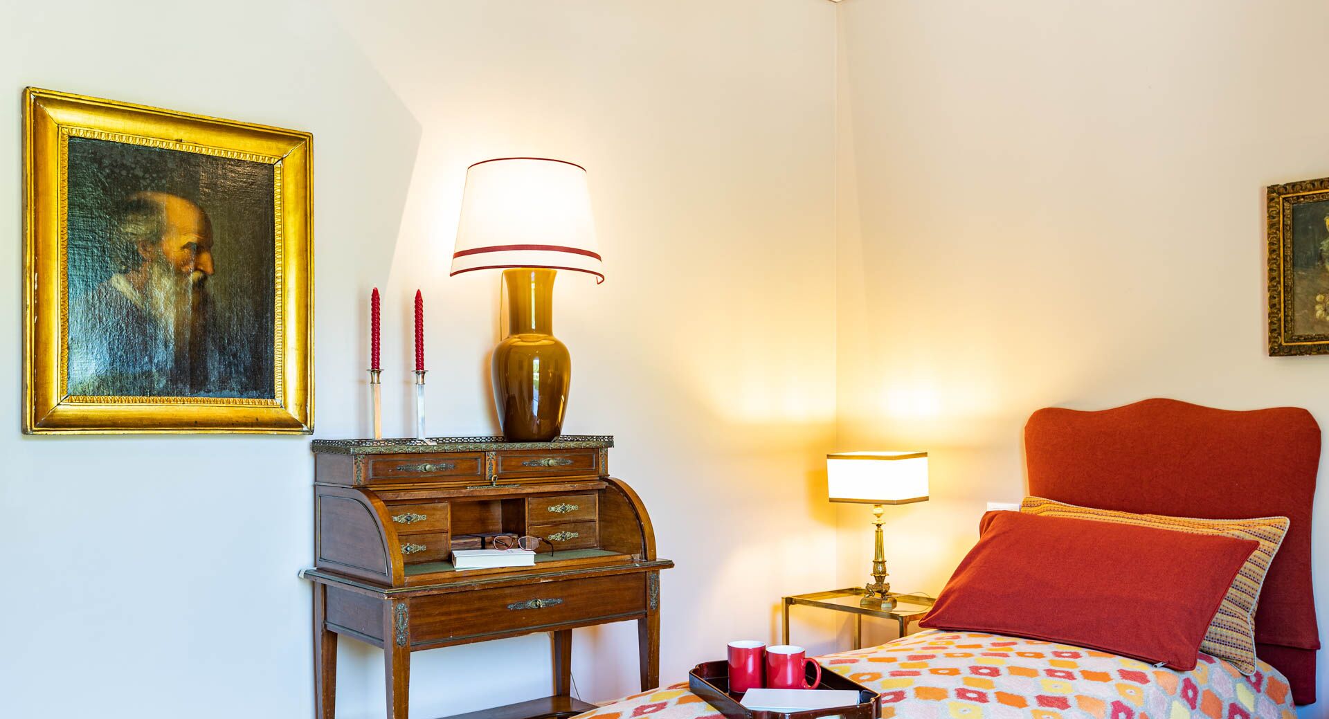 A hotel room with a wooden dresser, a lamp, a bed with red bedding, and two framed pictures on the wall.