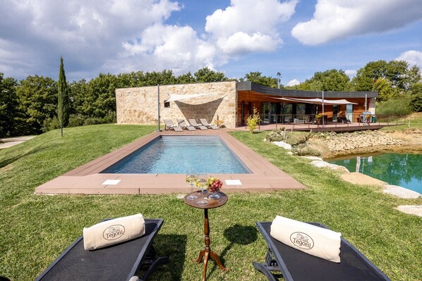 Un área de piscina moderna al aire libre con pared de piedra, sillas de descanso y una mesa con una copa de vino.