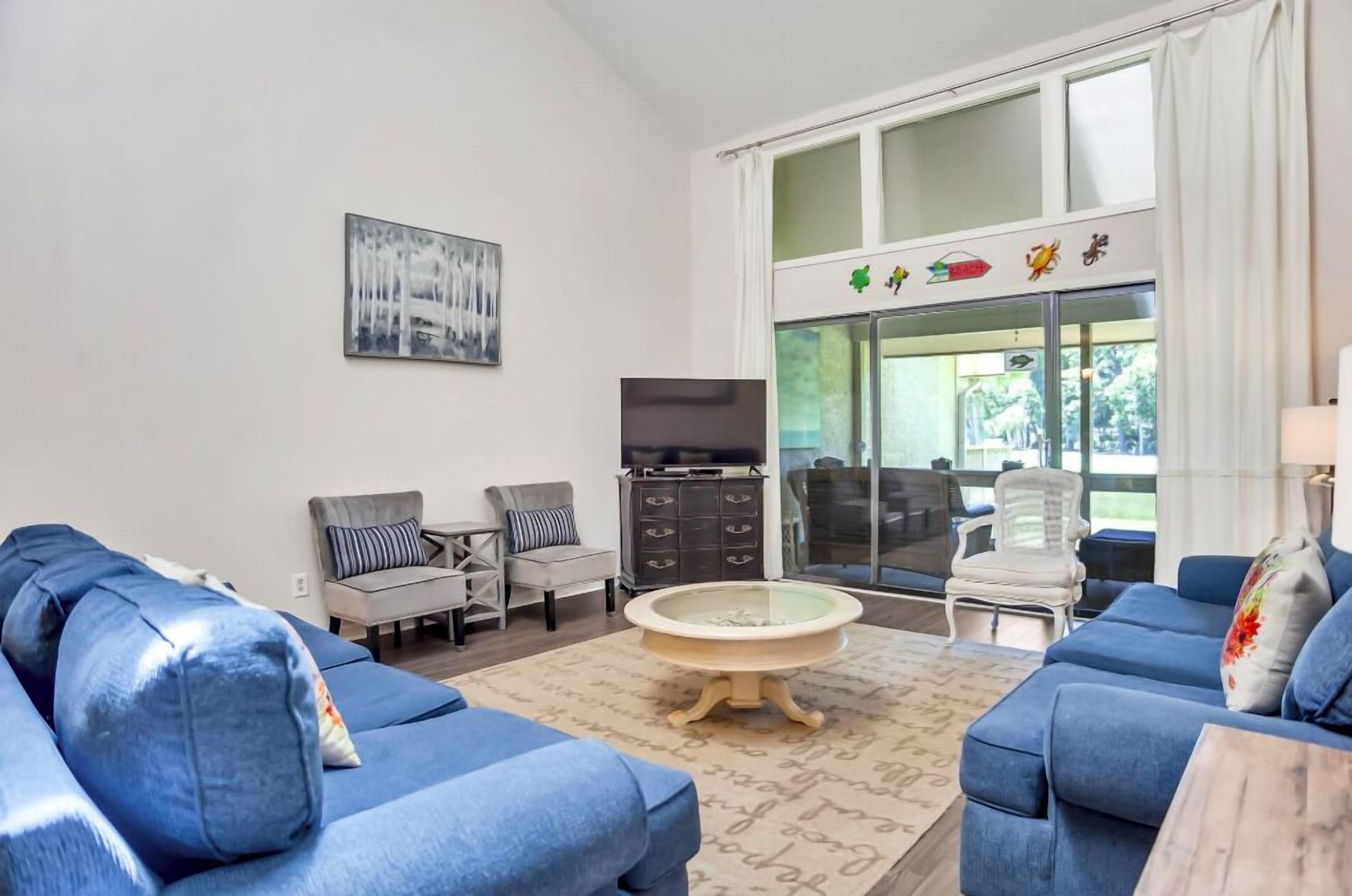 A living room with a blue sofa set, a round coffee table, a television, and a large window with a view of greenery.