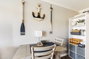Un comedor con una mesa de madera, dos sillas, un adorno de ancla colgado en la pared, un reloj de pared y un estante blanco con cajones.