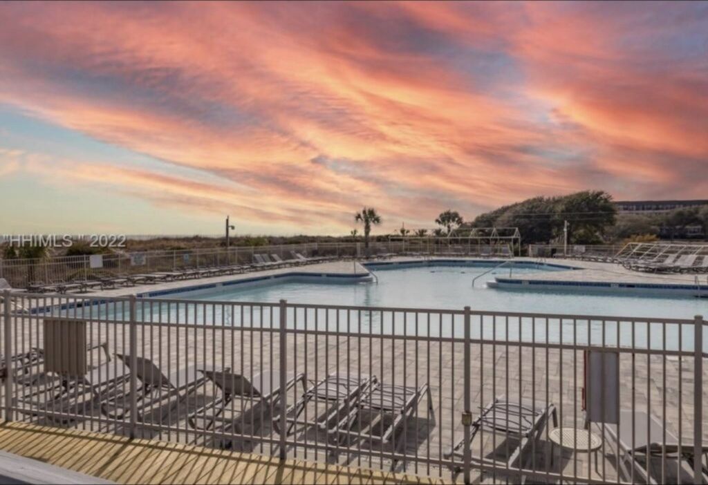 A swimming pool with lounge chairs and a fence, surrounded by trees and a sunset sky.