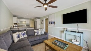 Un salon avec un canapé d’angle gris, une table basse en bois, un ventilateur de plafond et un téléviseur à écran plat fixé au mur.