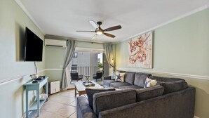 Un salon avec un canapé gris, une table basse, un téléviseur fixé au mur et un ventilateur de plafond.
