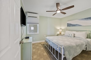 Un dormitorio con una cama, ventilador de techo, aire acondicionado, mesita de noche con lámpara y un cuadro en la pared.