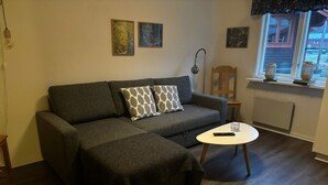 Ein Wohnzimmer mit grauem Sofa, zwei gemusterten Kissen, einem kleinen weißen Couchtisch und einer Stehlampe. An der Wand befinden sich gerahmte Bilder und ein Fenster mit Ausblick.