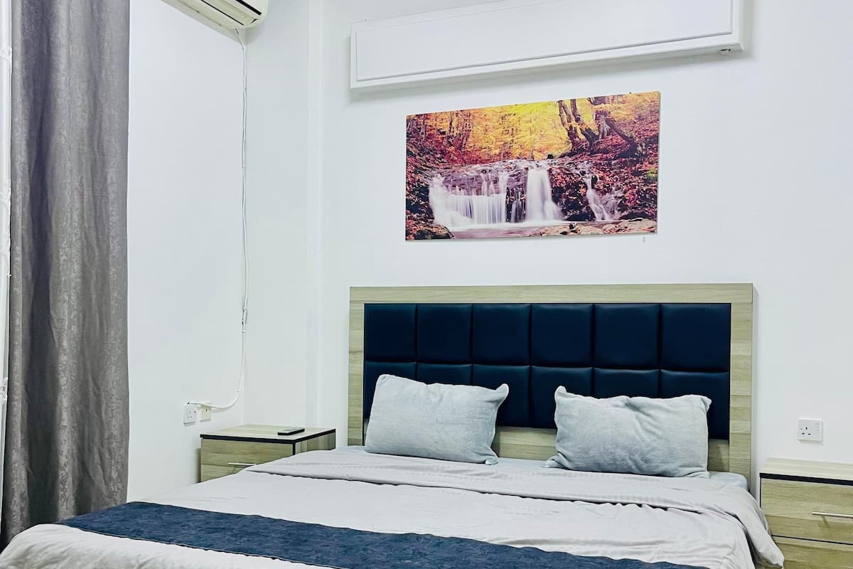 Un lit bien fait, avec une tête de lit, un climatiseur fixé au mur et un tableau représentant une cascade.