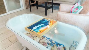 Een glazen salontafel met een kleurrijk schilderij en schelpen, een tafelvoetbaltafel en een bank met een bloemenkussen in een woonkamer.