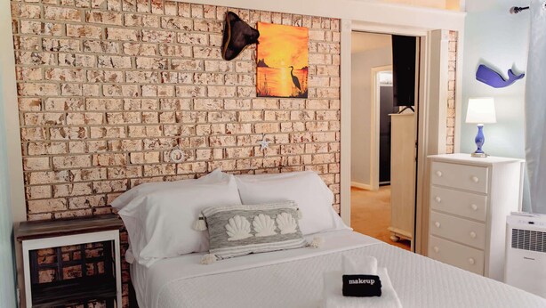Een slaapkamer met een wand in baksteenmotief, een bed met wit beddengoed, een decoratief kussen, een sterosier en een nachtkastje met een lamp.