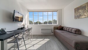 A modern, well-lit room with a large window, a flat-screen TV, a sofa, and a dining area with chairs. - Roland House - Deluxe Studio D (London)