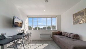 A modern, well-lit room with a large window, a flat-screen TV, a sofa, and a dining area with chairs. - Roland House - Deluxe Studio D (London)
