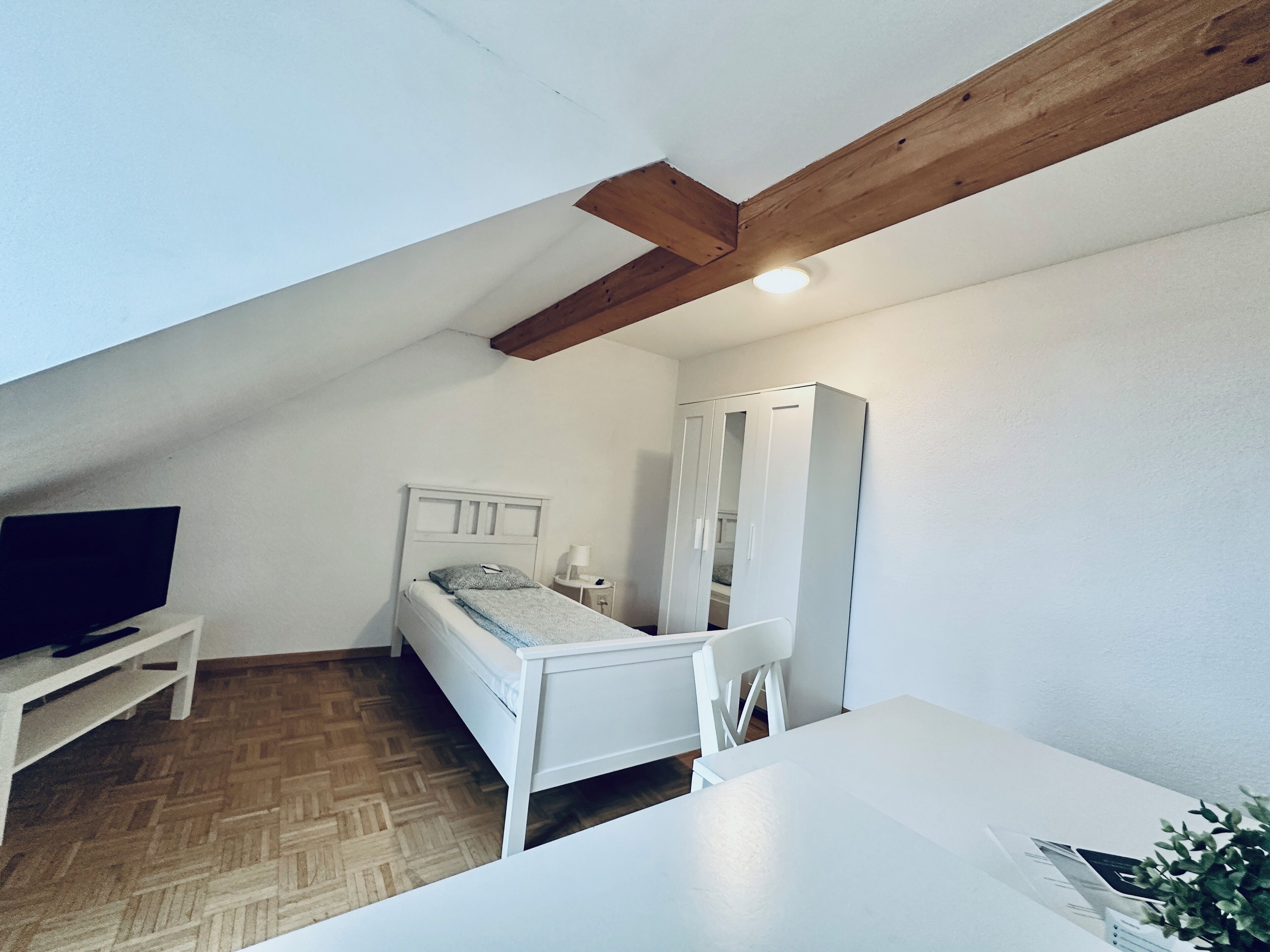 Un petit appartement d’une seule pièce, aux murs blancs, avec une poutre en bois au plafond, un lit, une armoire blanche, une petite table et un téléviseur posé sur un meuble blanc.