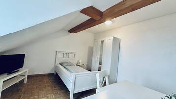 Un petit appartement d’une seule pièce, aux murs blancs, avec une poutre en bois au plafond, un lit, une armoire blanche, une petite table et un téléviseur posé sur un meuble blanc.
