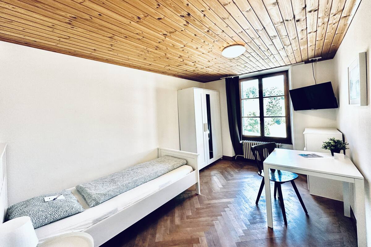 A compact room with a wooden ceiling, a white sofa, a small table, and a window with a view of trees.