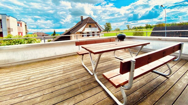 An outdoor wooden deck with picnic tables and a barbecue grill, overlooking a residential area and a grassy field under a partly cloudy sky. - ZIMMERzuVERMIETEN.ch Flumenthal SO (Flumenthal)