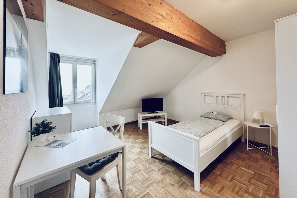 A bedroom with a sloped ceiling, a bed, a desk, a chair, a TV, and a window with curtains.