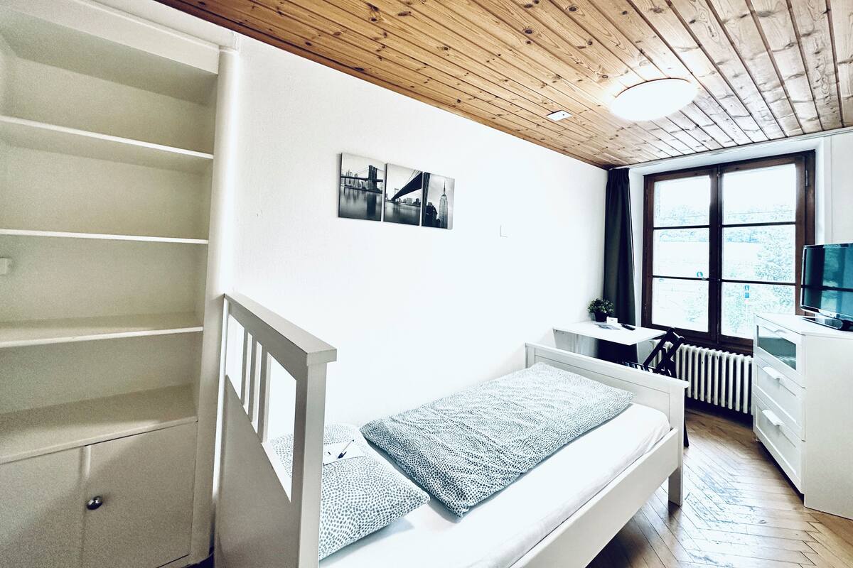 A bedroom with a wooden ceiling, a bed with a patterned blanket, a desk with a computer, and a window with a view of trees.