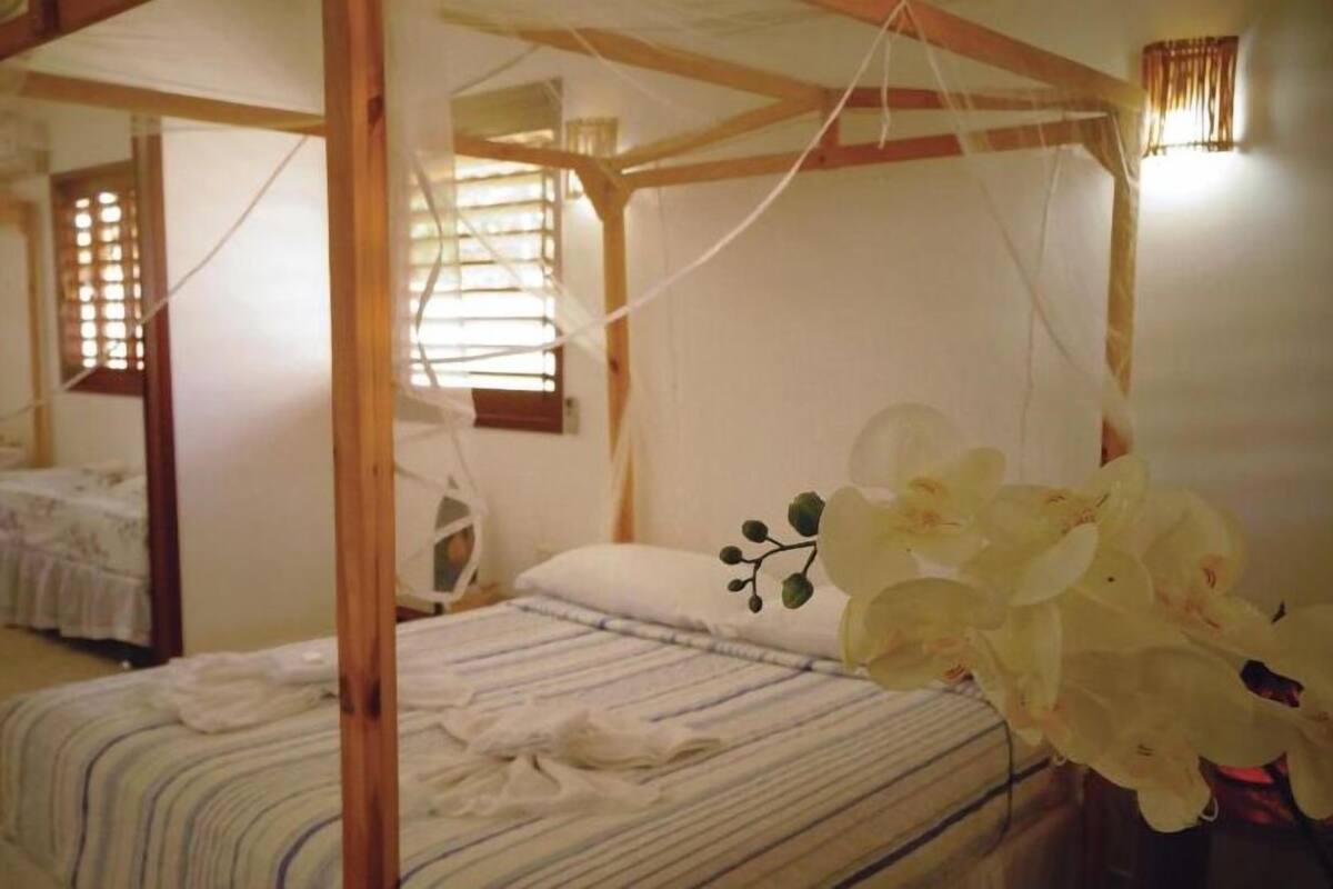 Una habitación con cama con dosel, una mesita y una silla. Se ve una ventana con persianas y cortina.