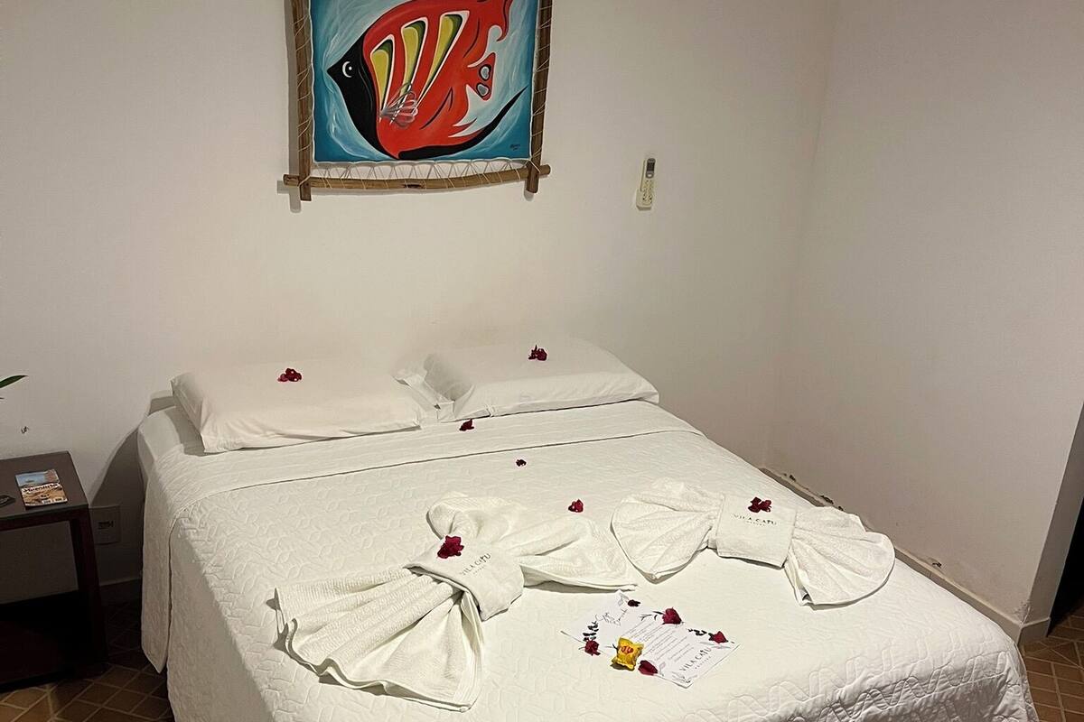 Una cama bien hecha con sábanas blancas y pétalos de rosa, un cuadro enmarcado encima y un suelo de baldosas.