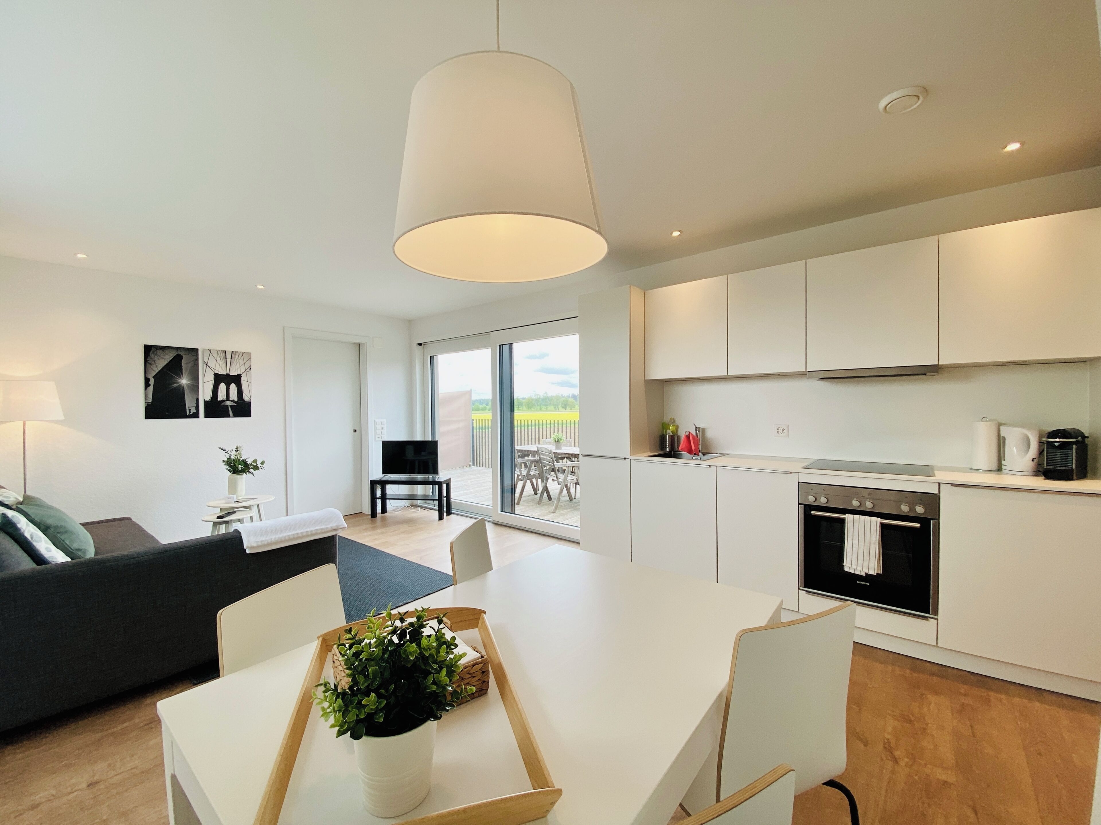 Una cocina moderna con armarios blancos, un comedor con mesa y sillas blancas, y un amplio ventanal con vistas al exterior.