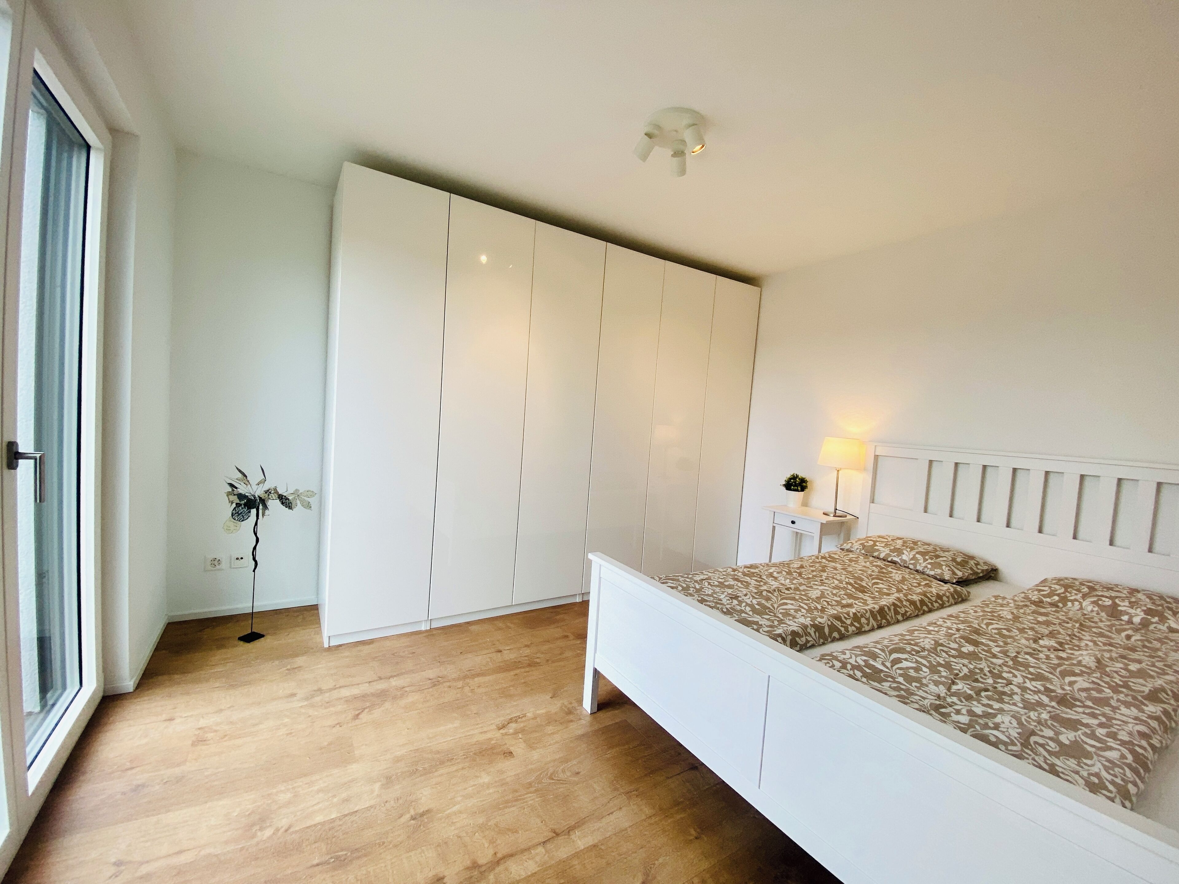 A bedroom with a bed, a wardrobe, a bedside table with a lamp, and a view of the outdoors.