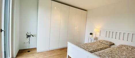A bedroom with a bed, a wardrobe, a bedside table with a lamp, and a view of the outdoors.