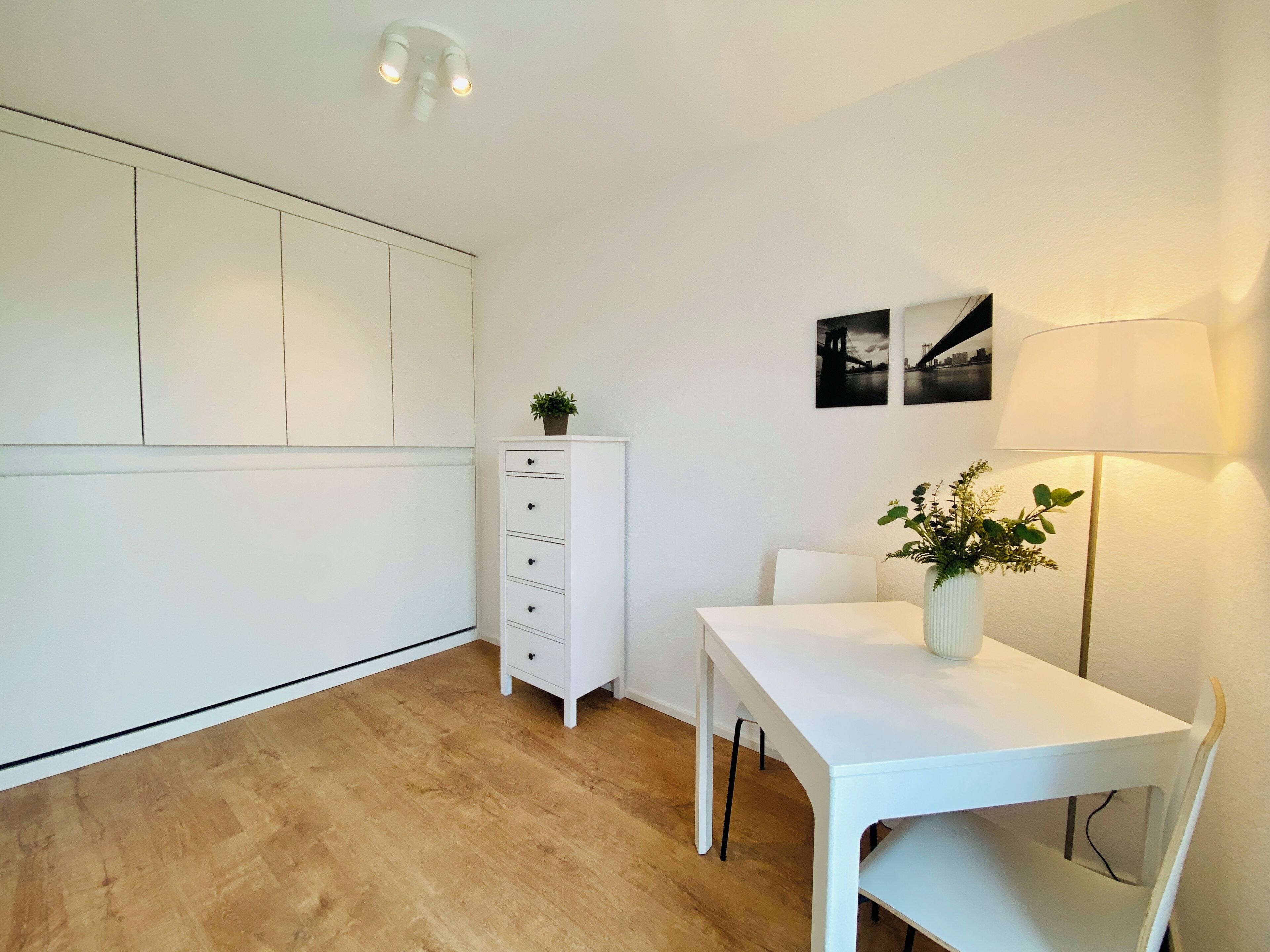 Une pièce moderne et minimaliste, avec une table blanche, un meuble blanc et un lampadaire. Deux tableaux encadrés ornent le mur.