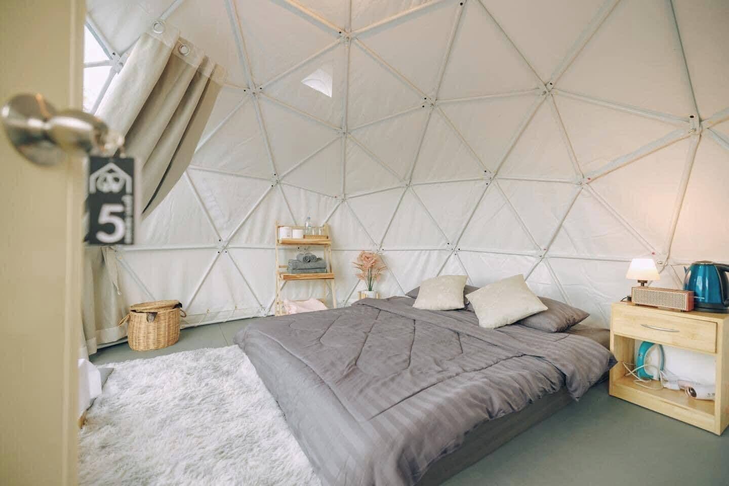 A tented room with a bed, bedside table, lamp, and a small shelf with toiletries.
