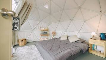 A tented room with a bed, bedside table, lamp, and a small shelf with toiletries.