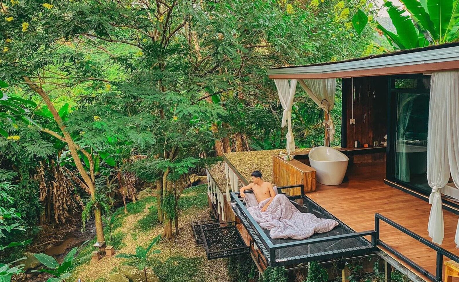 Un salon extérieur privé doté d’une terrasse en bois, d’un espace salon avec des coussins et d’une pièce fermée par des parois de verre, où se trouve une baignoire.