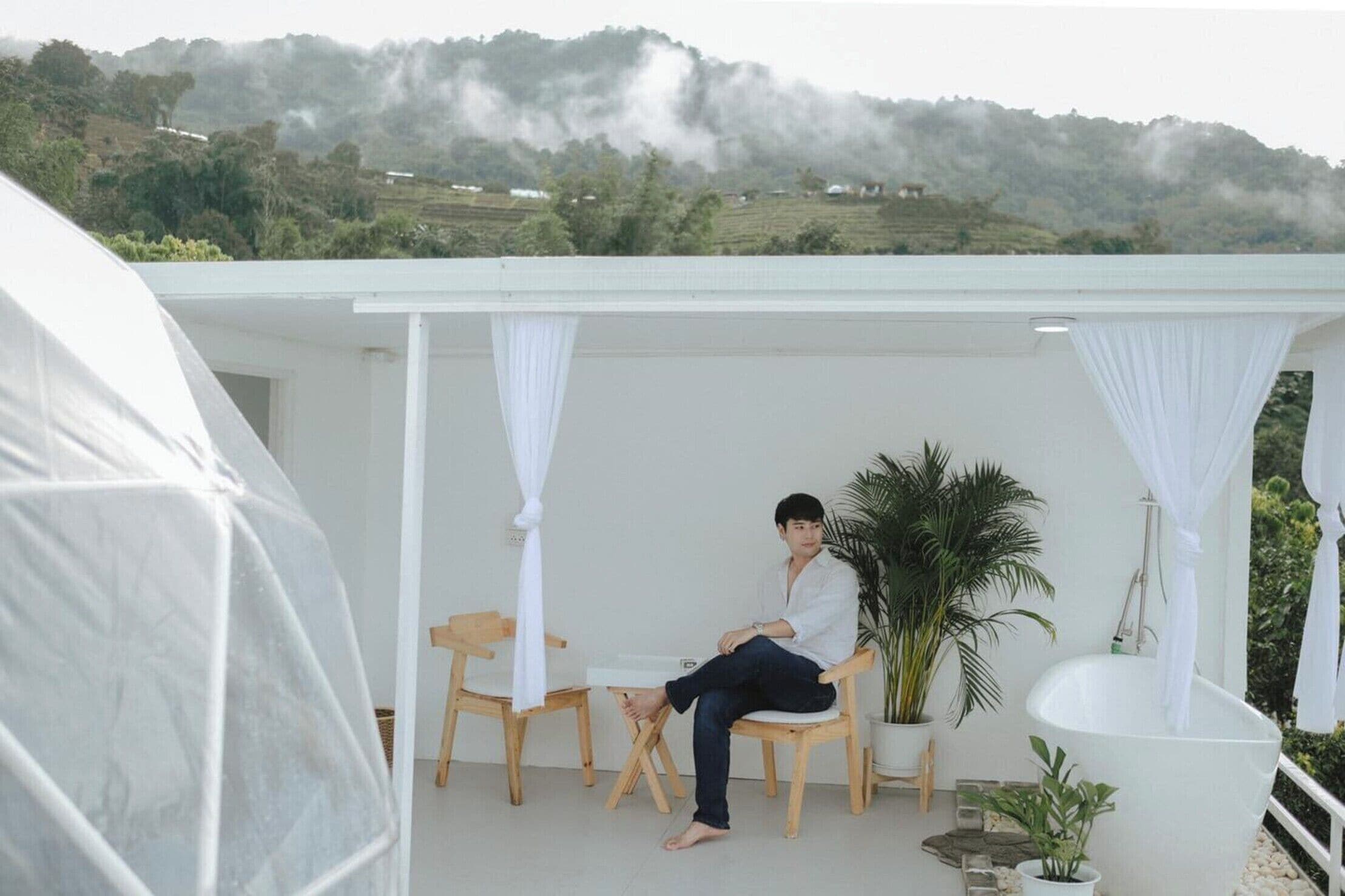 A man sitting on a chair under a white canopy on a balcony with a view of green hills and a white bathtub.