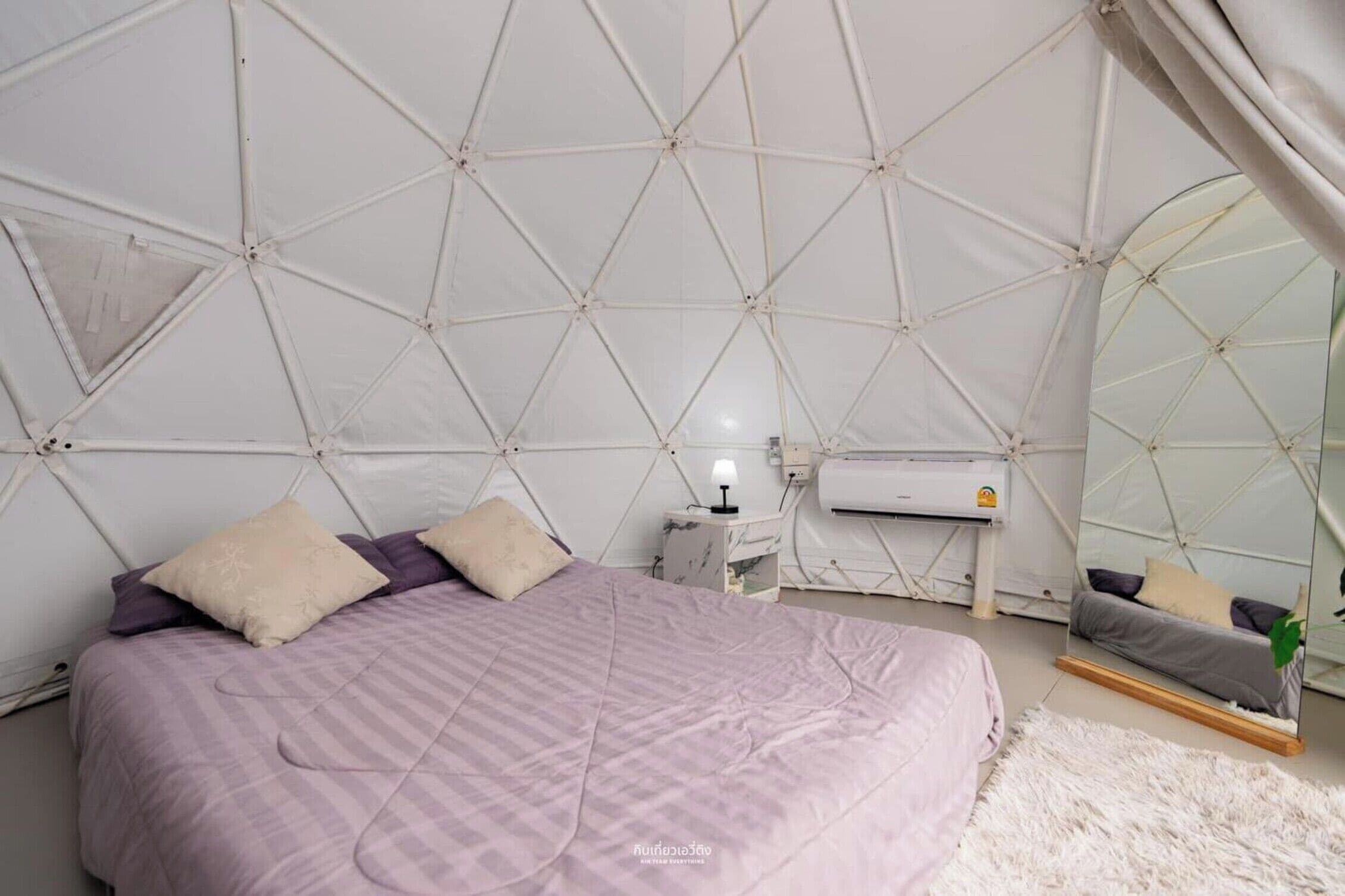 Ett sovrum med ett geometriskt mönster i taket, en säng med ett lila överkast, ett sängbord med en lampa och en spegel som reflekterar sängen.