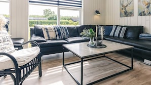 A modern living room with a black leather sofa, a glass coffee table, and a view of the outdoors through large windows. - 5 Star Holiday Home in Hadsund (Hadsund)