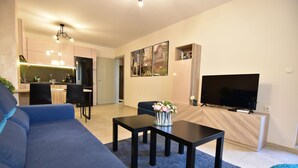 Una sala de estar moderna con un sofá, una mesa de centro y un televisor. Se ve una cocina al fondo con un comedor.