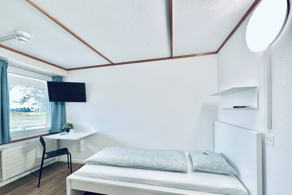 Een kamer met een bed, een bureau, een stoel, een televisie en een raam met gordijnen.
