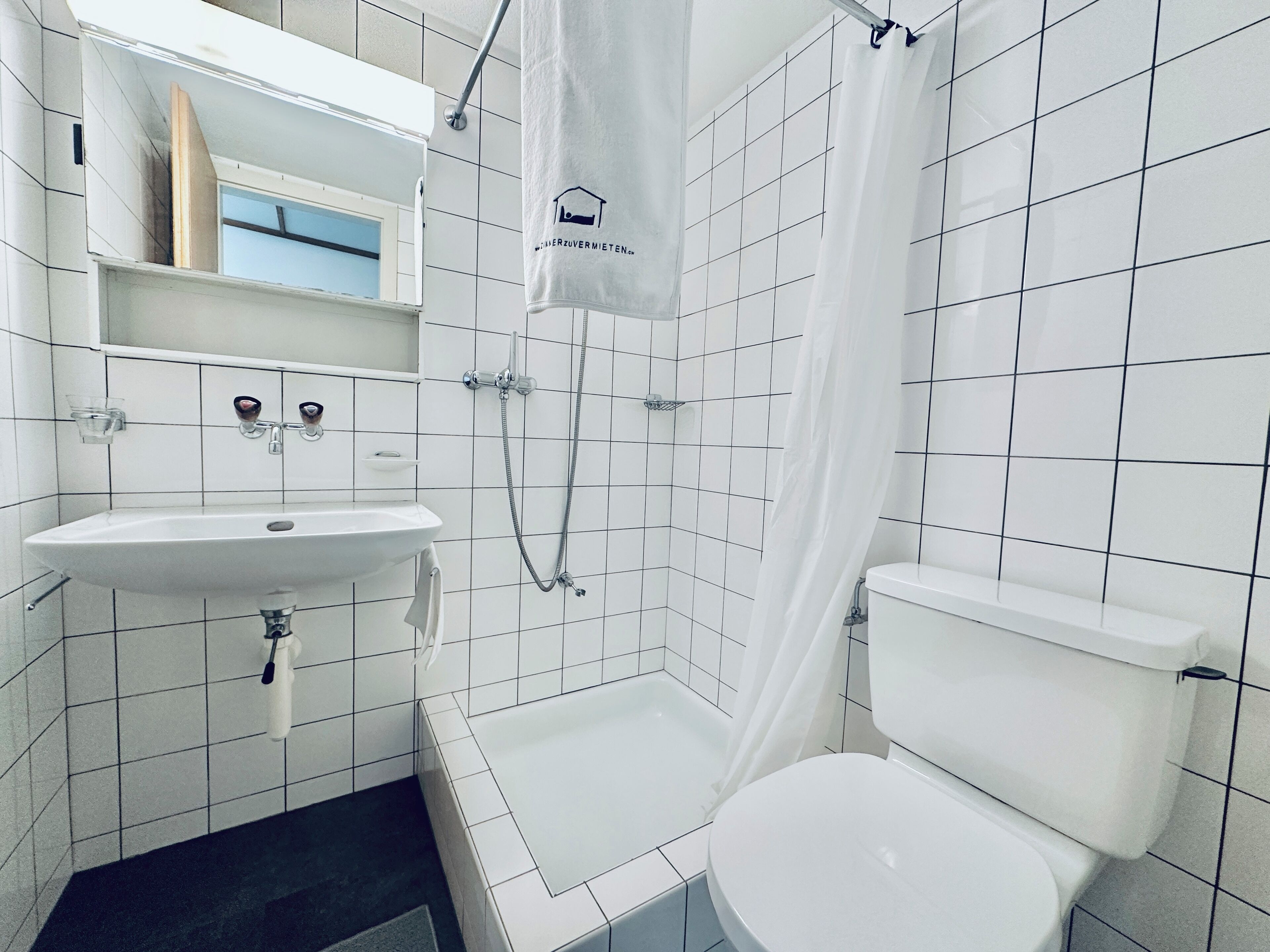 Un baño con lavamanos, inodoro y ducha blancos. Hay una toalla colgada en la cortina de la ducha.