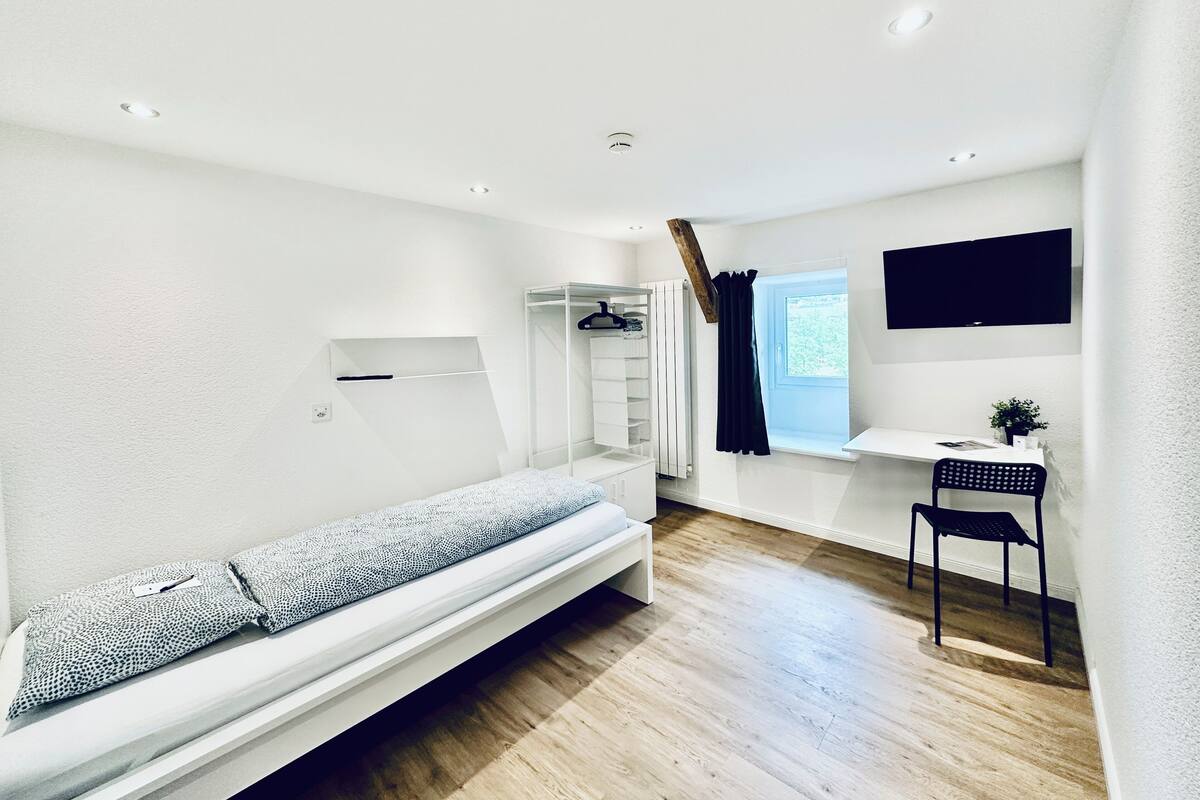 Een kleine, moderne kamer met een stapelbed, een bureau met stoel, een radiator en een raam met gordijnen.
