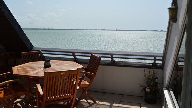 A balcony with a wooden table and chairs, overlooking a calm body of water.