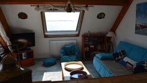 A cozy attic room with a large window, a blue sofa, a bean bag chair, a small table, and a bookshelf.