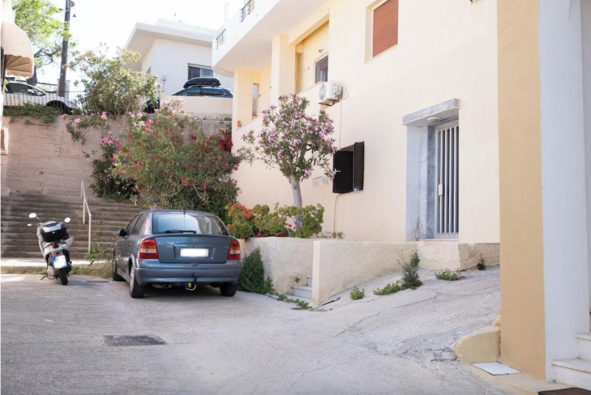 Rua estreita com um carro estacionado e uma motocicleta, ladeada por edifícios com varandas e pequenos jardins.